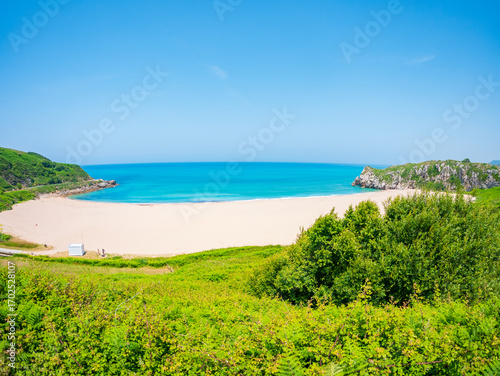 Panoramic view of Playa de Usgo in Cantabria, Spain, featuring turquoise waters, white sand, green cliffs, and a bright blue sky captured on a clear summer day.