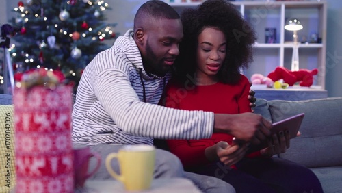 NEW YORK - December,13 - 2017. Pretty curly girl in red sweater sitting on sofa with boyfriend on background of wonderful tree. Girl using tablet. Hugging.