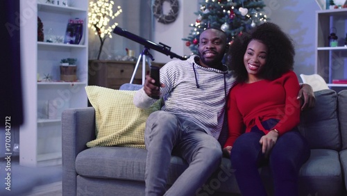 Happy African couple in love having rest in evening. Young people sitting on couch and viewing TV on background of decorated Christmas tree.