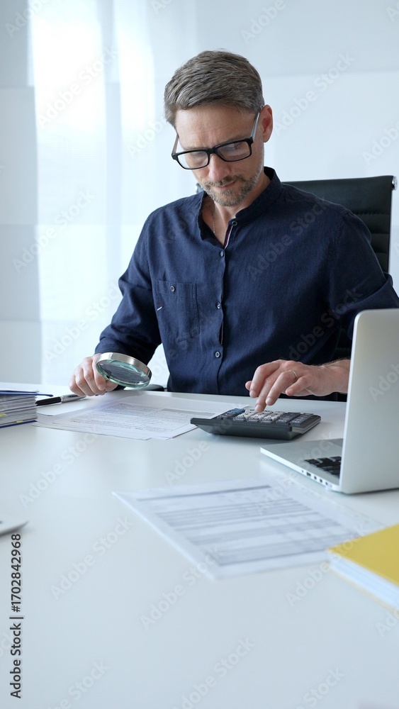 Fototapeta premium Businessman working in office set is using calculator and magnifying glass analyzing financial documents, performing accounting and financial analysis. Audit and taxes concept