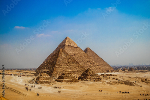 The three Great Pyramids of Giza rise above the golden desert, with long camel caravans and visitors crossing the sandy plateau while the skyline of Cairo is faintly visible on the horizon.