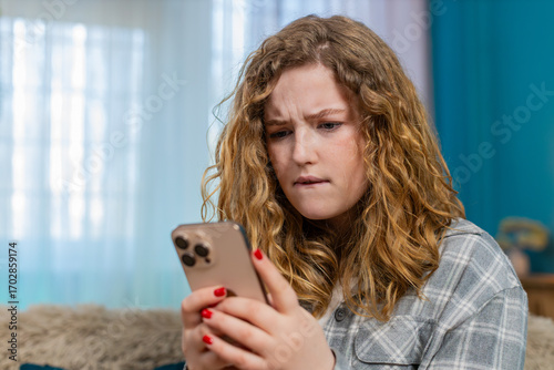 Young woman at home angrily texting on mobile phone reacting with irritation and frustration to message. Caucasian redhead girl on sofa waves hand annoyed, emotional expression of dissatisfaction