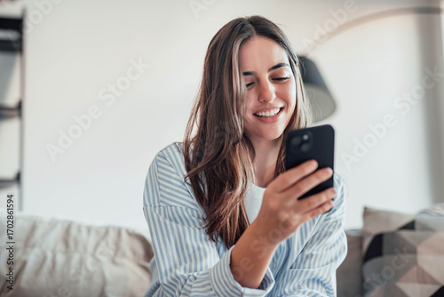 Photography Smiling woman looking at smartphone screen, sitting on couch, enjoying leisure t