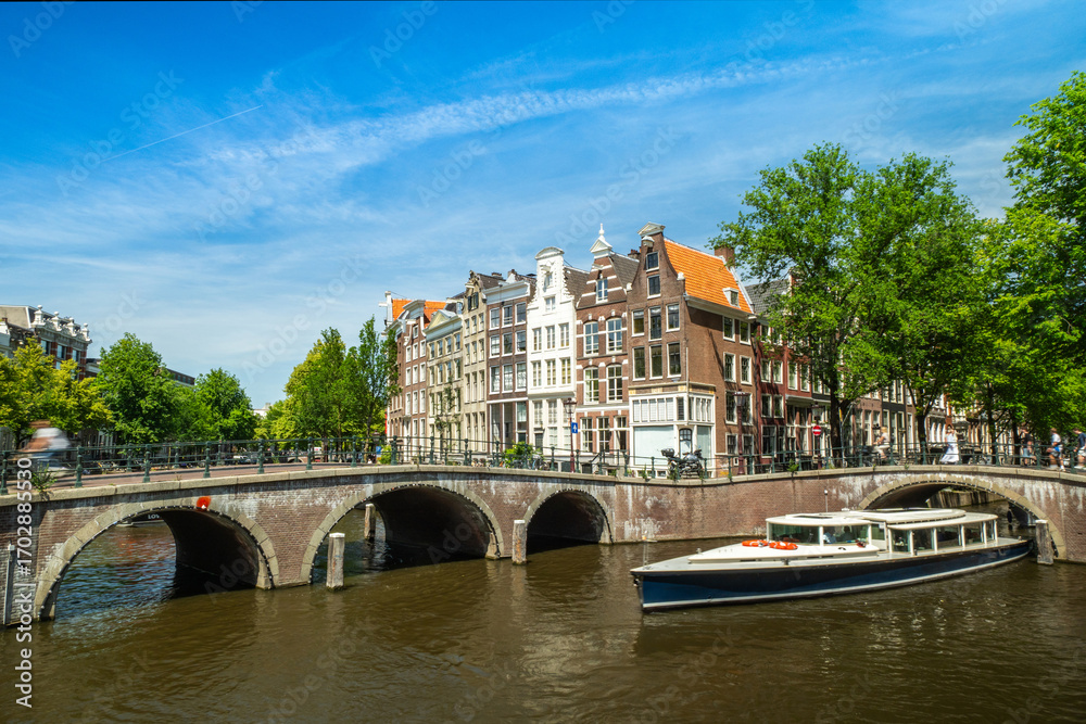Naklejka premium Beautiful canal view with historic Dutch architecture, traditional bridge, and tourist boat under clear blue sky in Amsterdam, Netherlands, perfect for travel and tourism concepts.