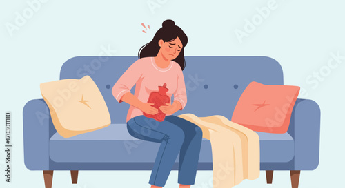 A woman in a pink shirt and blue jeans sits on a blue couch holding a red hot water bottle on her abdomen