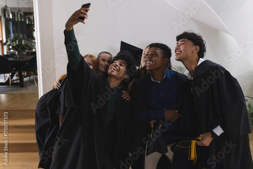 Graduating teens in caps and gowns taking selfie, celebrating together indoors