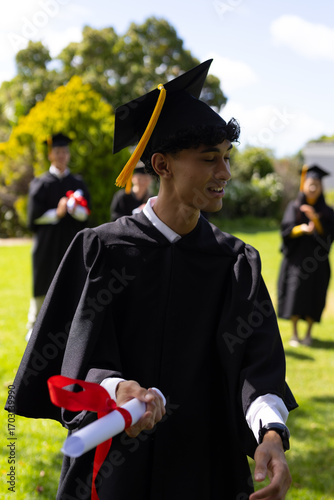 Teen graduate holding diploma, smiling outdoors with classmates in background
