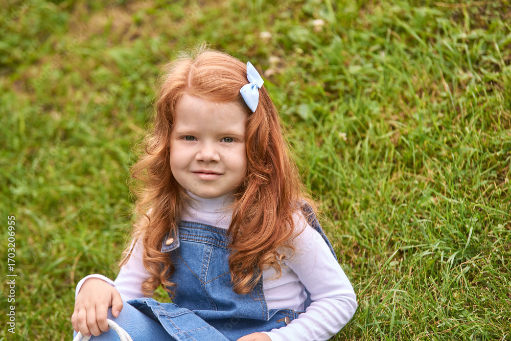 Fototapeta premium Red-haired caucasian young girl in denim dress sitting on grass with blue bow smiling.