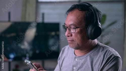 Mature man enjoys digital content. He wears headphones, holds smartphone, browsing media. Experience leisure, entertainment, communication using modern gadgets. Relaxed individual connects world.