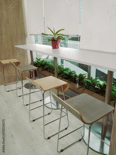 Minimalist interior design with wooden bar stools, tall white table, indoor plants, and city view outside the window. Bright and modern office space concept.