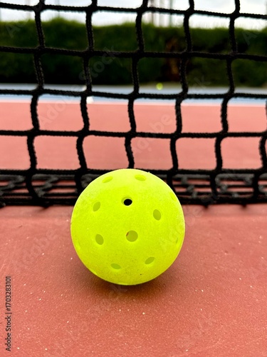 Pickleball ball close up on the hard surface by the net.  Yellow perforated, hollow plastic ball for pickleball sport 