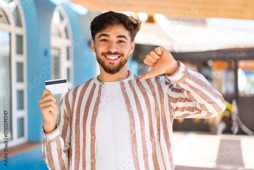 Handsome Arab man holding a credit card at outdoors proud and self-satisfied
