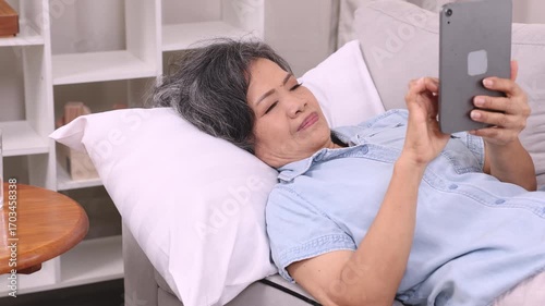 Mature Asian woman lying on bed and holding smartphone, enjoying technology at home in peaceful solitude. This relaxed lifestyle shows senior, woman, phone, home, technology, independent living.