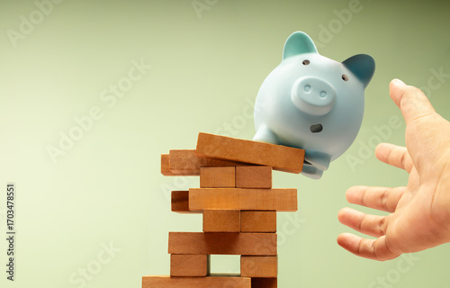 Fotografía Piggy bank suddenly falls off unstable Jenga block, hands protecting it, symboli