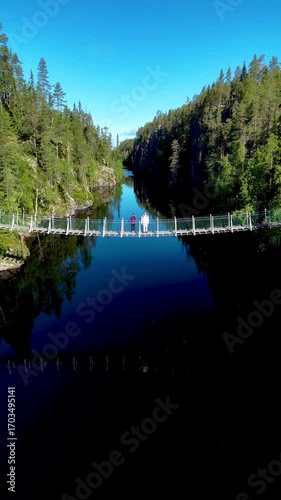 Visitors experience the serene beauty of Canyon Lake Julma Olkky, surrounded by lush forests and calm waters in Hossa National Park, Finland. A couple walks at the suspension bridge over the canyon