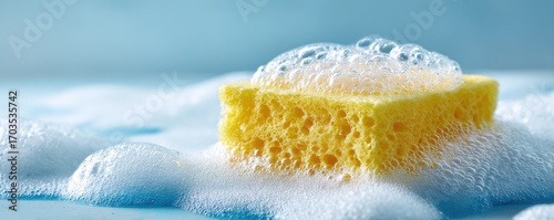 A yellow cleaning sponge sits in soapy bubbles on a light blue surface