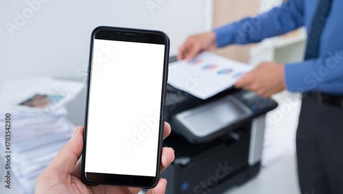 A hand holds a smartphone with a blank white screen in an office as a man uses a multifunction printer in the background.
