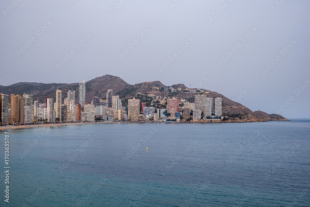 Fototapeta premium Benidorm, Spain, Coastal city skyline featuring modern architecture along the shoreline, with tranquil waters reflecting the buildings and a distant mountainous backdrop, creating a urban landscape