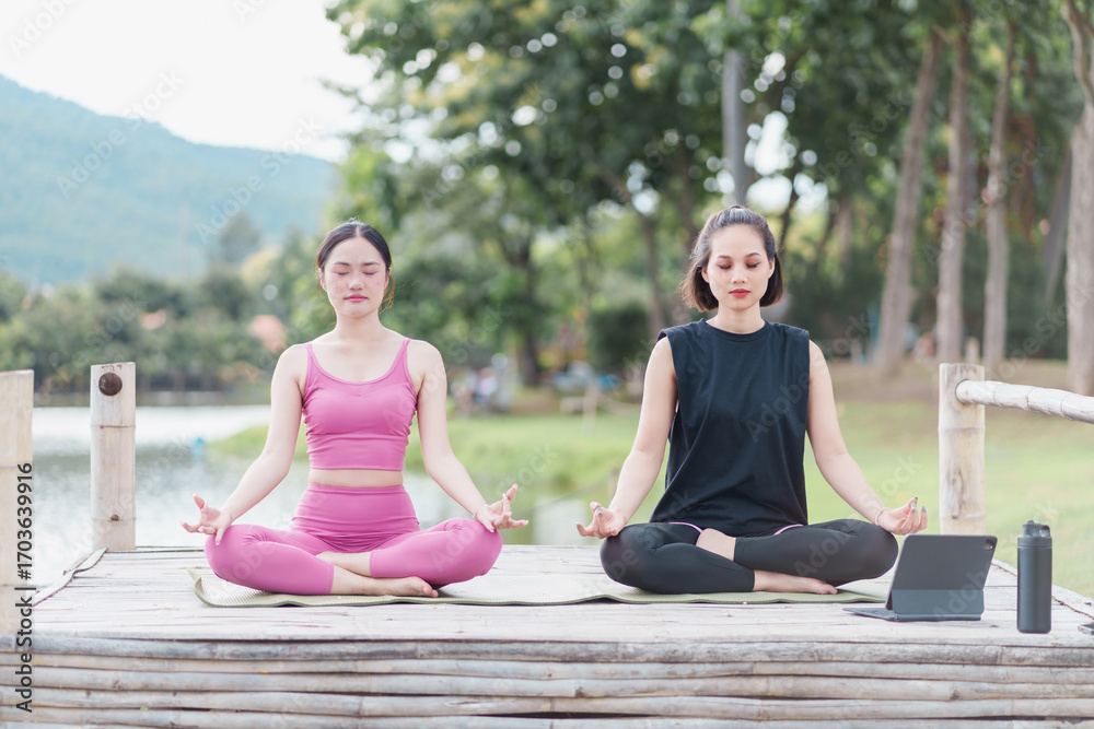 Fototapeta premium Young woman yoga meditation by lake in park, serene outdoor mindfulness practice with tablet and water bottle