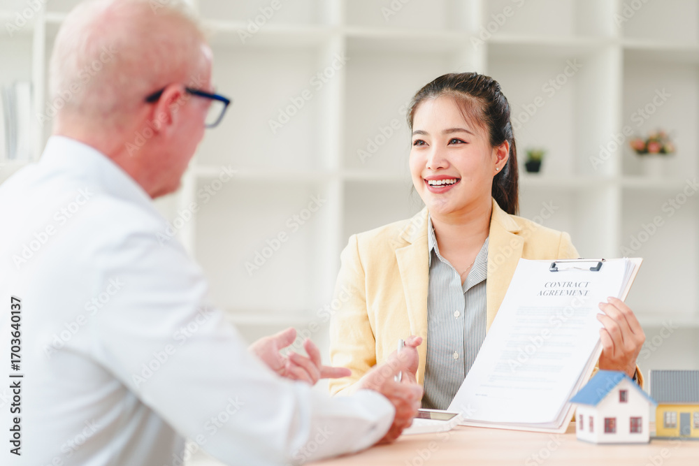 Fototapeta premium Young real estate agent smiling while presenting contract agreement and house model at meeting, confident professional discussing terms warmly