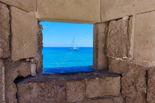 Sea view from ancient wall, travel and vacation concept. Turquoise sea with sailboat, summer holiday, stone window to the ocean sea.  Rhodes Island Greece, Fortress in the old town.