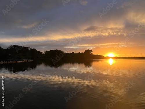 Summer sunset landscape sun setting over Norfolk Broads England uk tranquil water with the beautiful orange yellow blue skies viewed from sailing boat to horizon and trees reeds, clouds reflected 