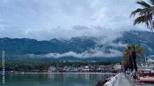 Scenic view of Akyaka, Turkey, with misty mountains, palm trees, and waterfront promenade. Atmospheric cloudy landscape with seaside town, nature, and coastal beauty.