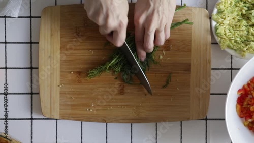 Hands cutting green dill with sharp knife, close up of cooking process, preparation of herbs for healthy salad, vegan food, and homemade dishes.