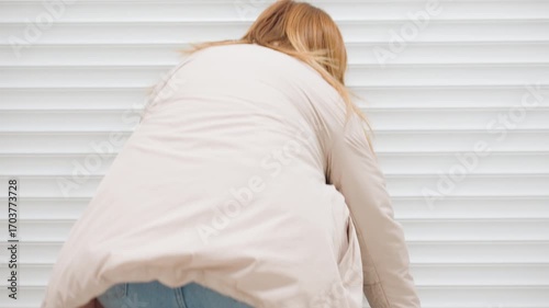 Back view of owner operator bends to roll up steel shutter of laundry room, hair moving, light jacket, motion blur, indoor corridor setting, clean space, morning routine, equipment inside dim