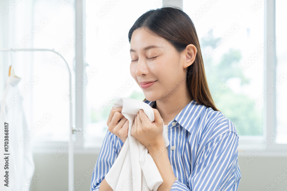 Fototapeta premium Feel softness, hygiene. Smile asian young woman touching fluffy white shirt smelling fresh clean clothes, pretty girl comfort sniff after washing laundry. Household work at home, chore of maid concept