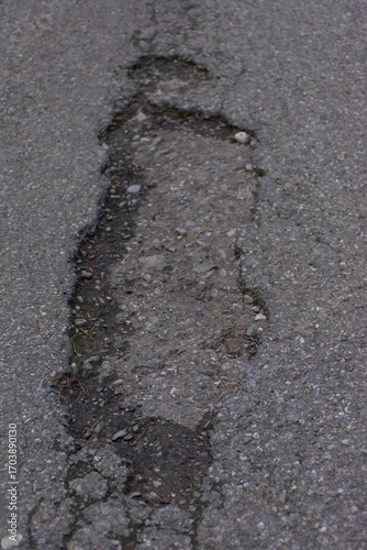 A cracked and weathered asphalt road with visible gravel and a single fallen leaf, creating a textured and moody image of urban decay, damage, and neglect.