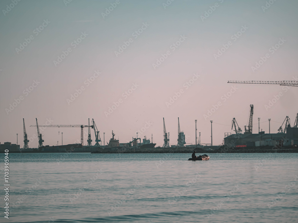 Obraz premium Tranquil harbor photo of a small boat on calm waters with a distant industrial port and cranes under a soft sky. Perfect for travel decor, maritime art, and eco-friendly designs.