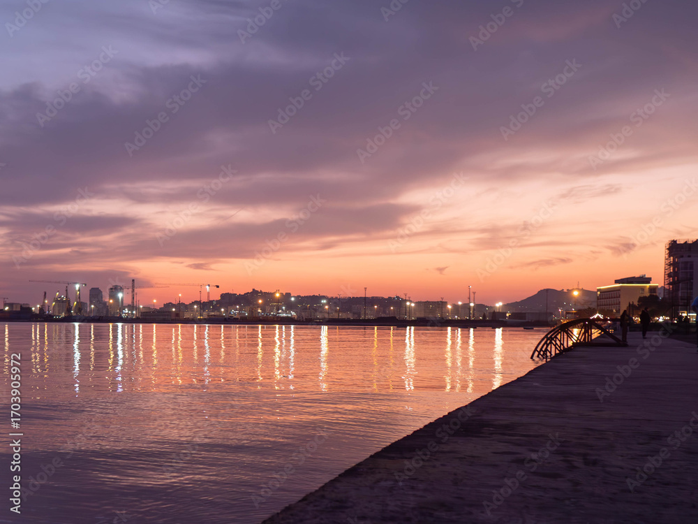 Fototapeta premium Breathtaking sunset photo of a city skyline reflecting on calm waters, with a serene promenade in the foreground. Ideal for travel decor, coastal art, and eco-friendly designs.
