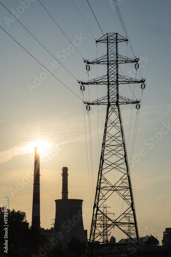 High voltage power lines and industrial plant with chimneys at sunset, symbolizing importance of reliable electricity infrastructure, energy distribution, technology, and sustainable urban development
