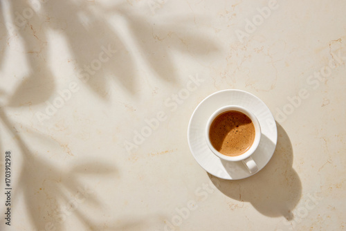 Fresh espresso served in a white cup and saucer on a beige stone surface, highlighted by natural light.