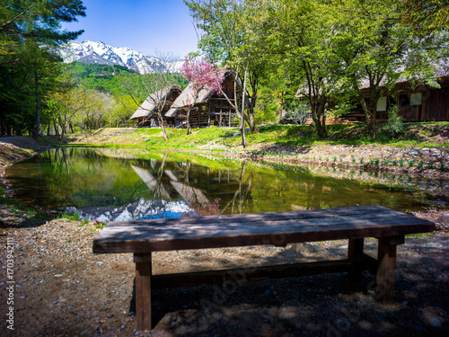 Scenic view of traditional thatched roof houses with a pond and snow-covered mountains in the background, in rural Japan.