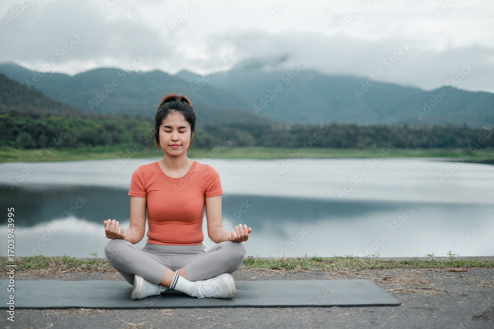 Fototapeta premium A woman practices meditation by a tranquil lake with mountains in the background, promoting relaxation and mindfulness.