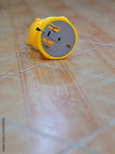 Yellow plastic toy, shaped like an animal, lies on a tiled floor with a wood-like pattern. The toy's base features a battery compartment with screws and a switch, typical in battery-operated devices.