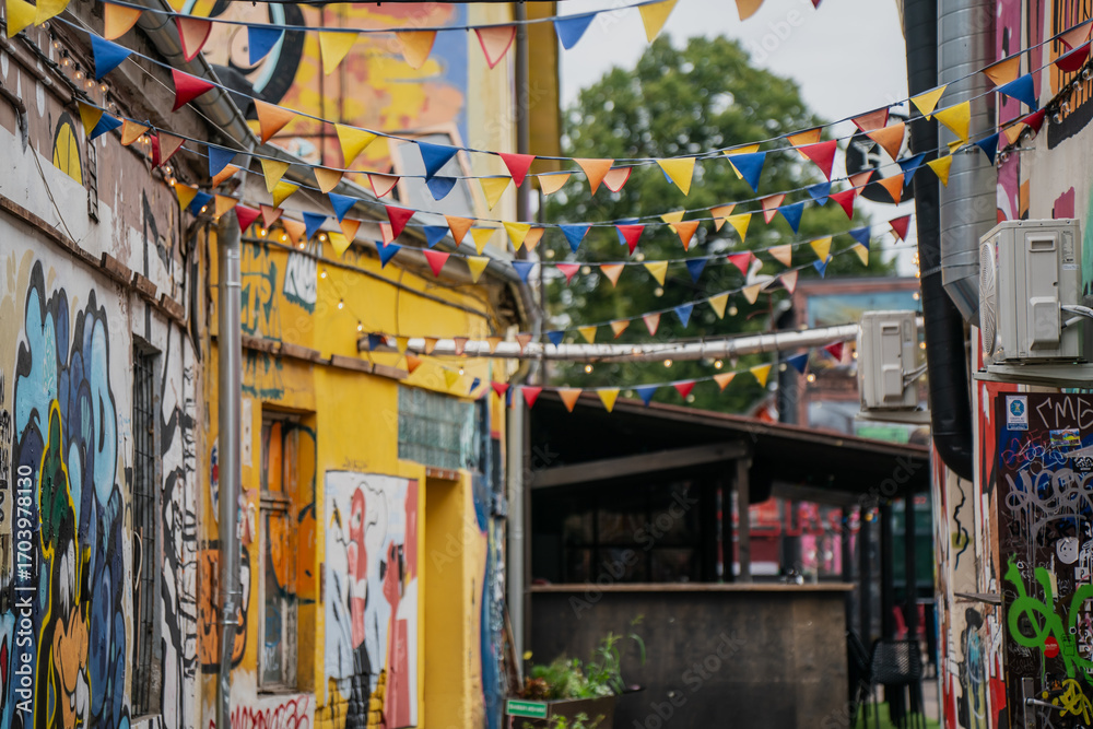 Obraz premium Urban alley decorated with colorful triangular pennant flags hanging between graffiti walls and murals, outdoor street scene featuring creative artistic expression and festive atmosphere