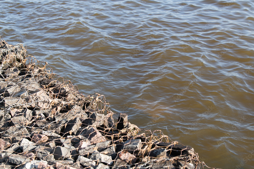 Obraz premium Water with a brownish tint softly touches a rocky shoreline, where stones are secured with a netting material, creating a tranquil riverside setting