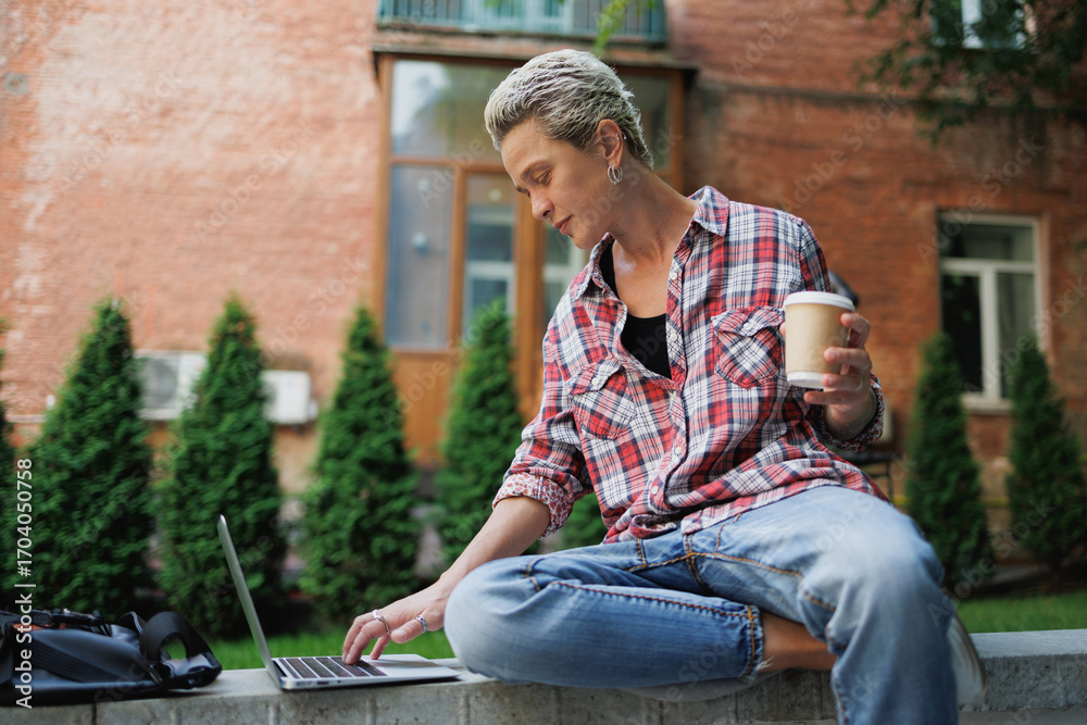 Obraz premium Stylish freelancer holding takeaway drink and using laptop outdoors 