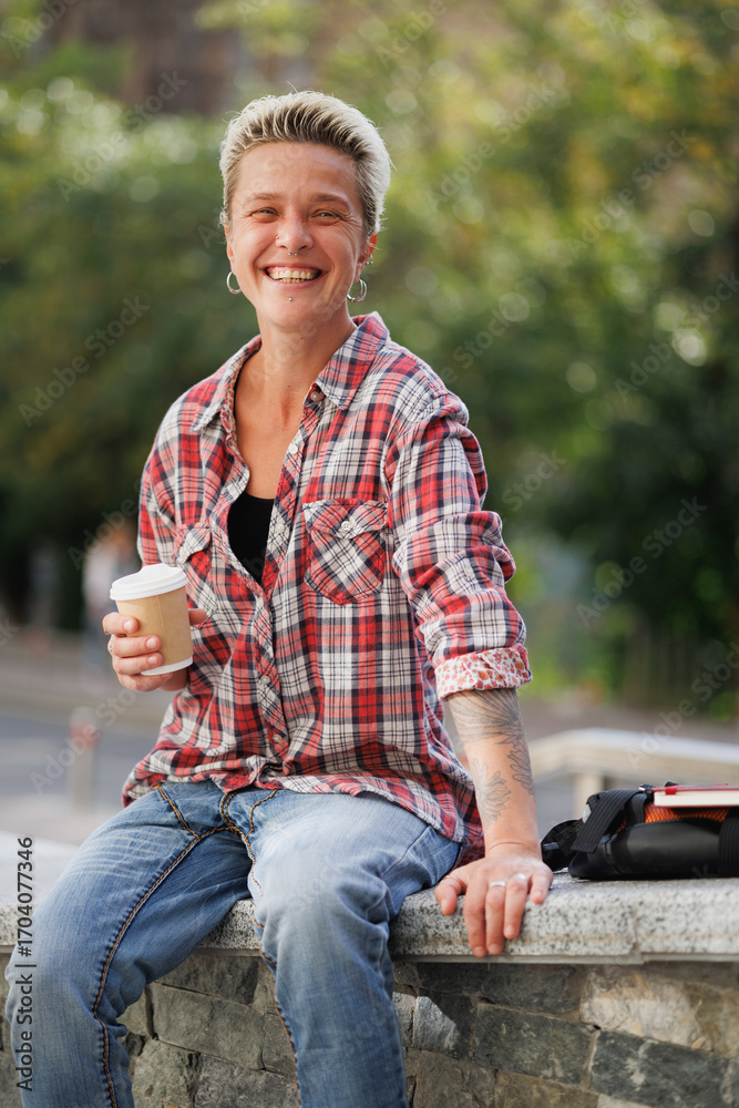 Obraz premium Cheerful tattooed woman holding coffee to go near bag and notebook on street