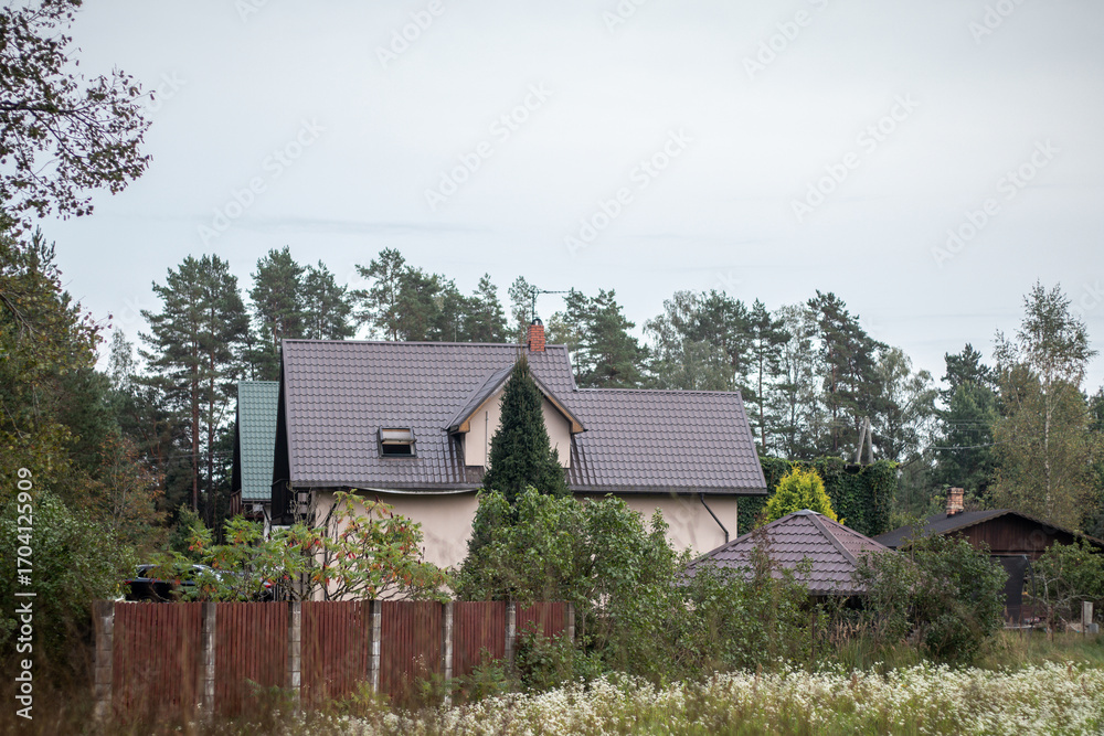 Fototapeta premium A beige suburban house with a dark tiled roof sits behind a wooden fence, surrounded by lush trees and vegetation under a cloudy sky.