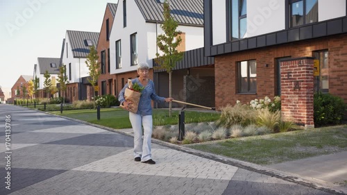 Older woman with short gray hair carrying grocery bag filled with vegetables and bread while holding cane. Female walking on quiet residential street near modern houses enjoying relaxed moment.