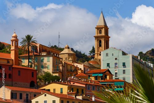 A view of a beautiful Italian old town Vintimille rising up into the hillside towards mountains in the distance. There are painted houses and church spire and wooded hills.