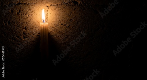A single lit candle casts a warm glow against a dark, textured wall.