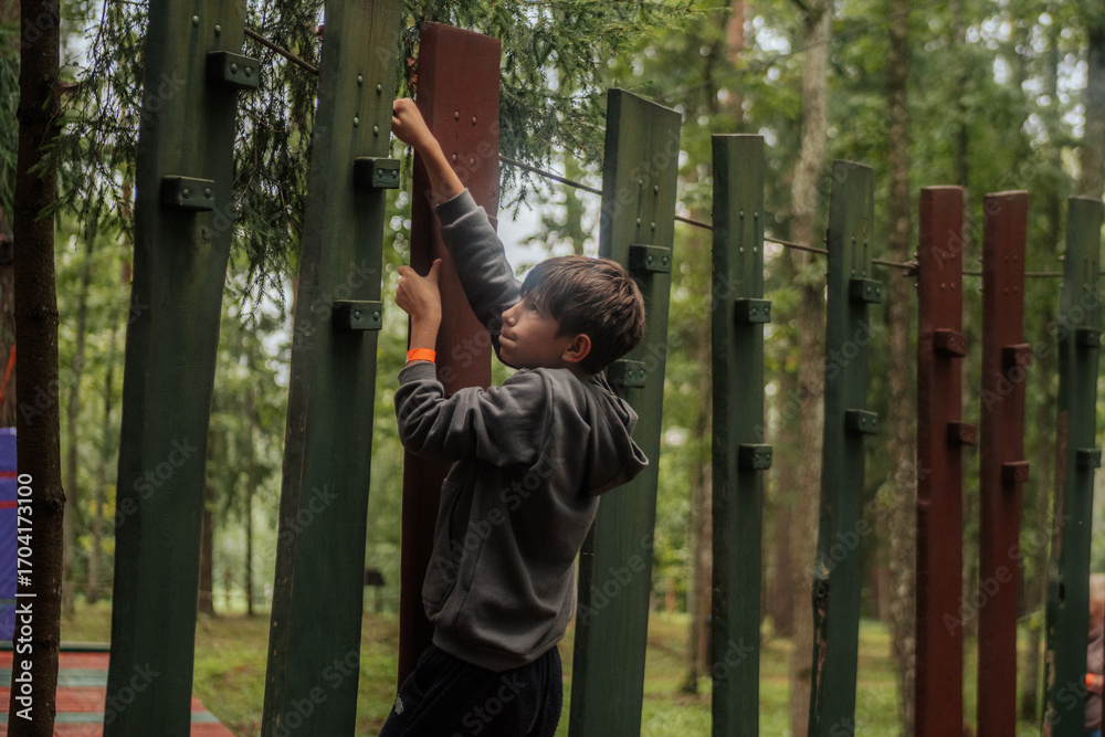 Obraz premium A focused boy in a hoodie climbs vertical wooden poles in an outdoor forest obstacle course, holding on tightly with both hands in a natural forest environment. 
