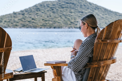 Beach workation digital nomad plans tasks with laptop and notebook on lounge chairs, powered by a portable solar charger. Eco-friendly, productive, flexible lifestyle by the sea. now