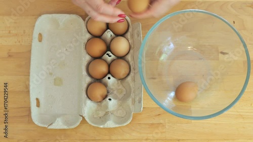 Person is cracking eggs into a bowl. The eggs are in a carton and the bowl is on a wooden table