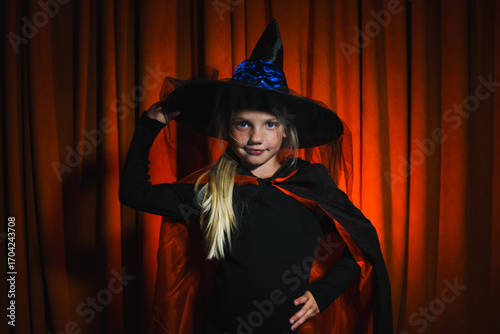 Joyful little witch posing for Halloween celebration in a spooky atmosphere with orange drapes and mystical flair